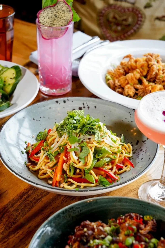 Vibrant noodle bowl, calamari, avocado salad, and pink drinks on table.
