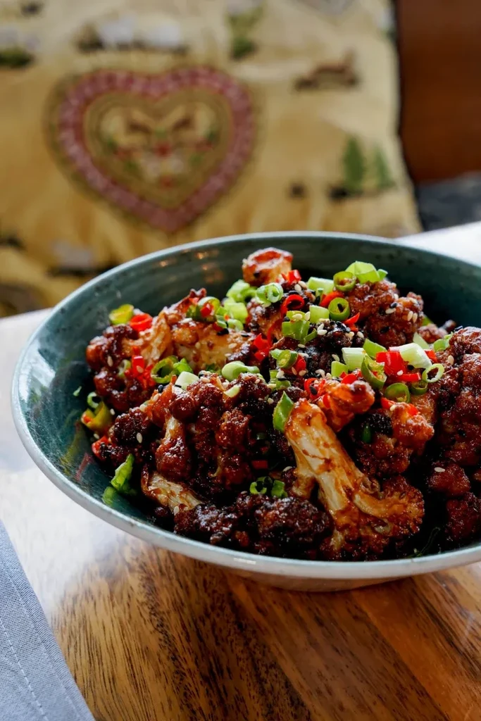 Roasted cauliflower with dark sauce, scallions, chili, and sesame in teal bowl.