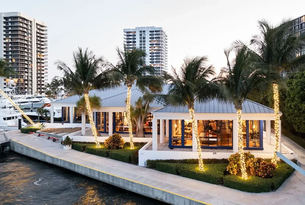 Modern white building by water with lit palm trees and yachts.