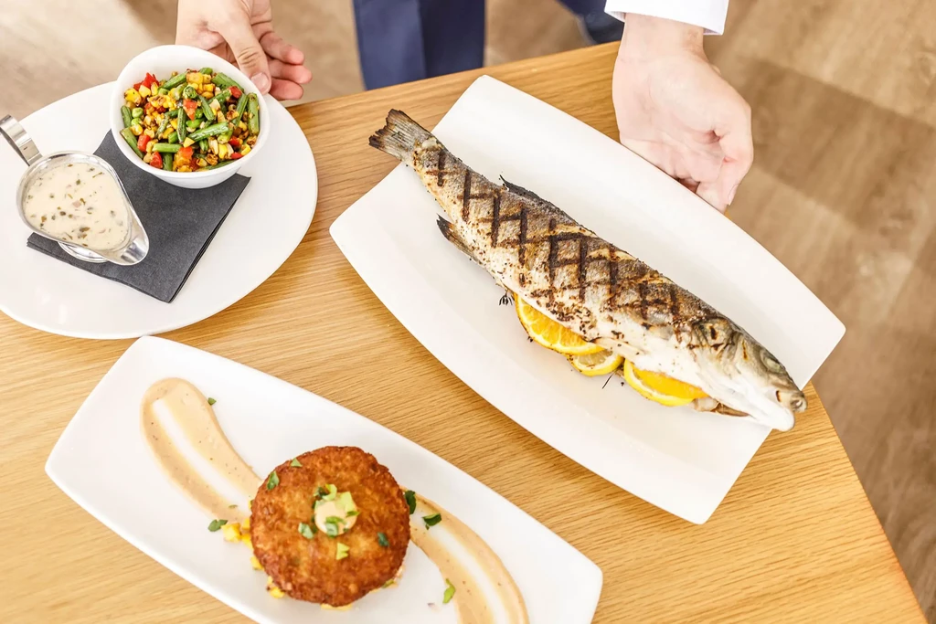 Top-down view of grilled fish, salad, and dishes presented by hands.