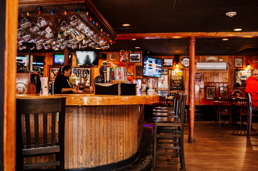 Cozy bar interior with wooden counter, stools, hanging mugs, and bartender.