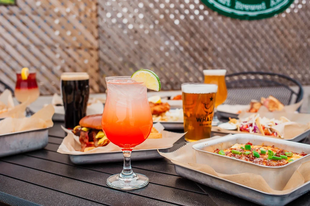 Vibrant layered cocktail, beers, burger, tacos, and cheesy dish on a patio.