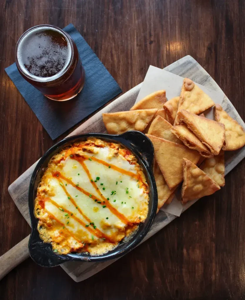 Baked cheesy dip, tortilla chips, and beer on a wooden table.