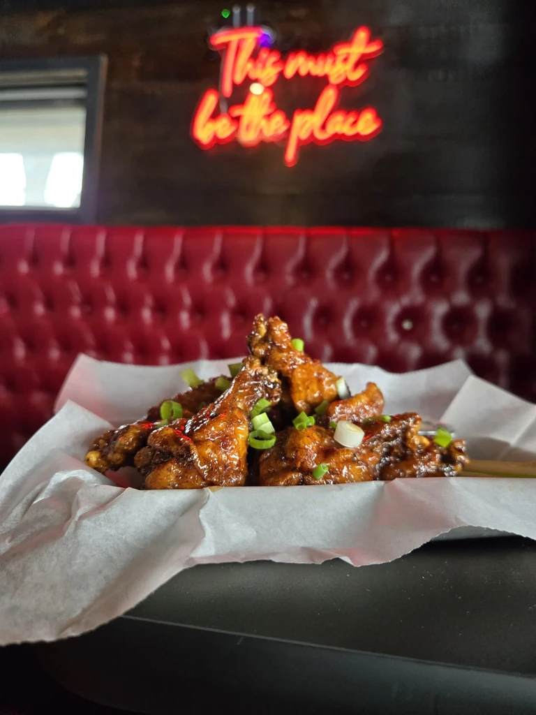 Basket of glazed chicken wings with onions and a red neon sign.