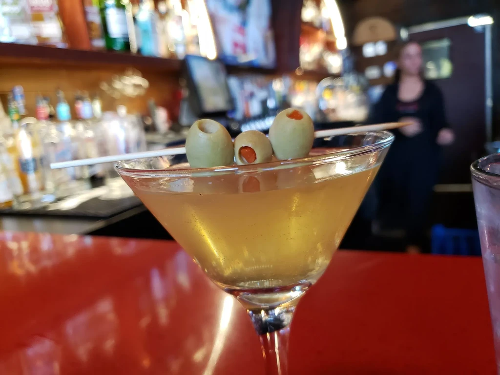 Martini with yellowish drink and three green olives on red bar.
