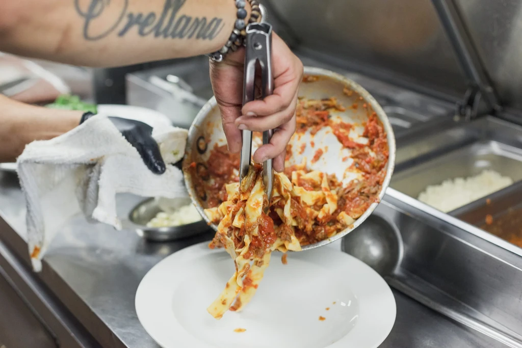 Chef's gloved hands use tongs to plate pasta with red sauce.