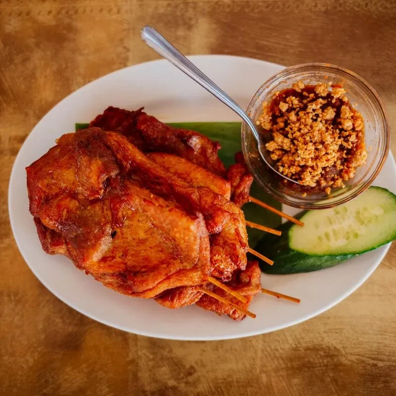 Skewered fried meat on banana leaf with brown sauce and cucumber.