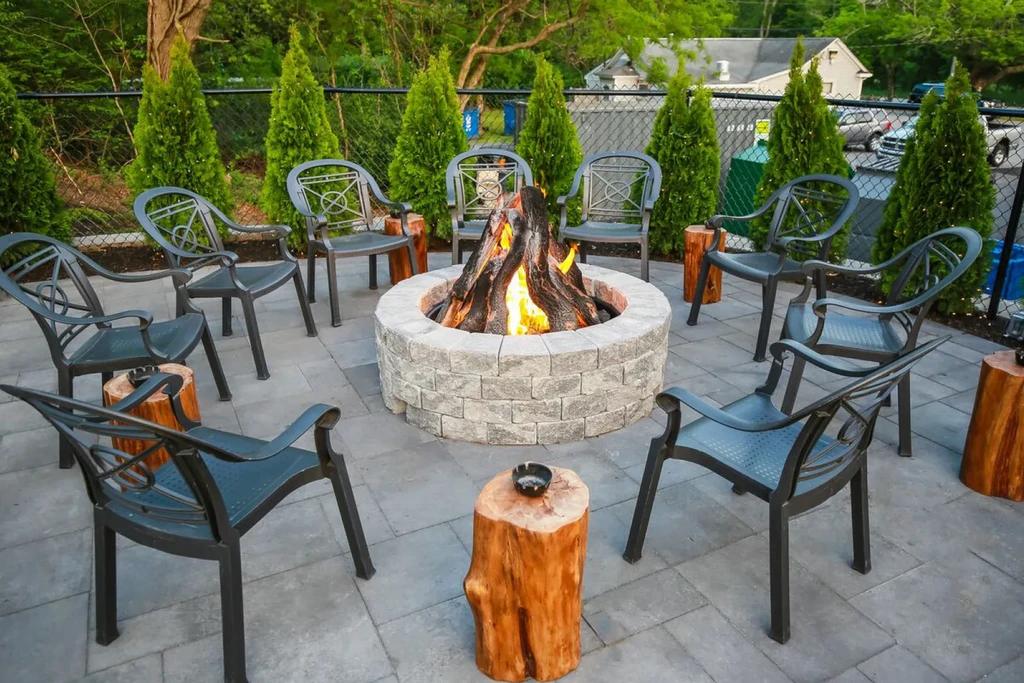 Paved outdoor patio with fire pit, seating, and evergreen shrubs.