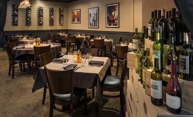 Cozy restaurant interior with warm lighting, set tables, and wine bottles.