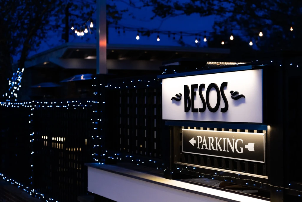 Illuminated "BESOS" restaurant sign with "PARKING" sign below at night.
