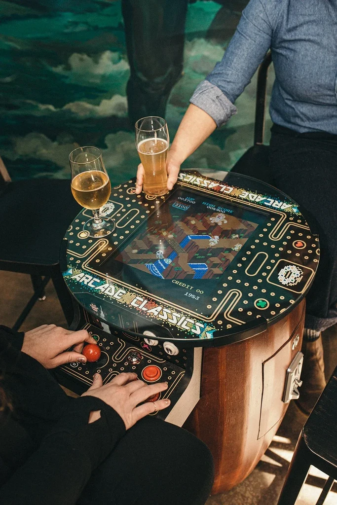Overhead view: two people play classic arcade game, beer nearby.
