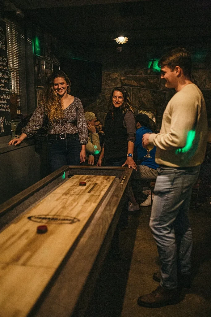Three smiling people play shuffleboard in a warm bar.