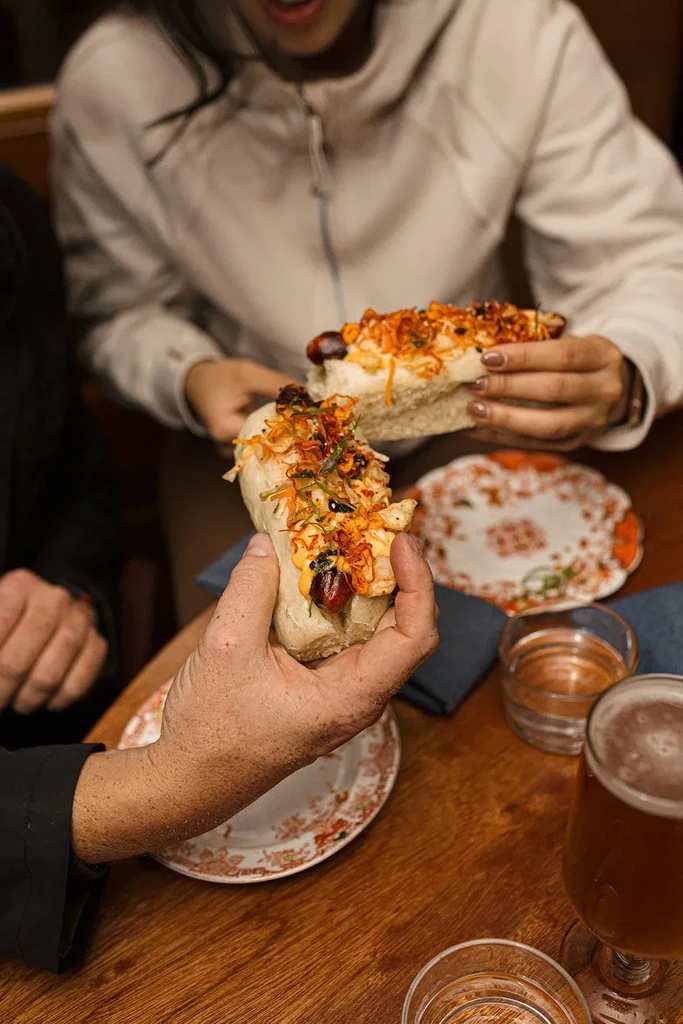 Two hands holding richly topped gourmet hot dogs on a rustic table.