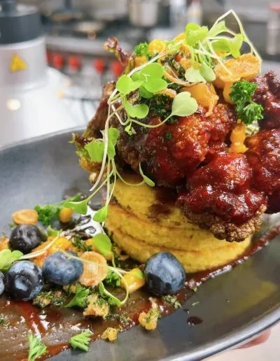 Plated stack of yellow fritters, crispy protein, microgreens, and blueberries.