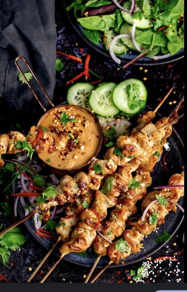 Satay chicken skewers, peanut sauce in copper pot, cucumber, salad.