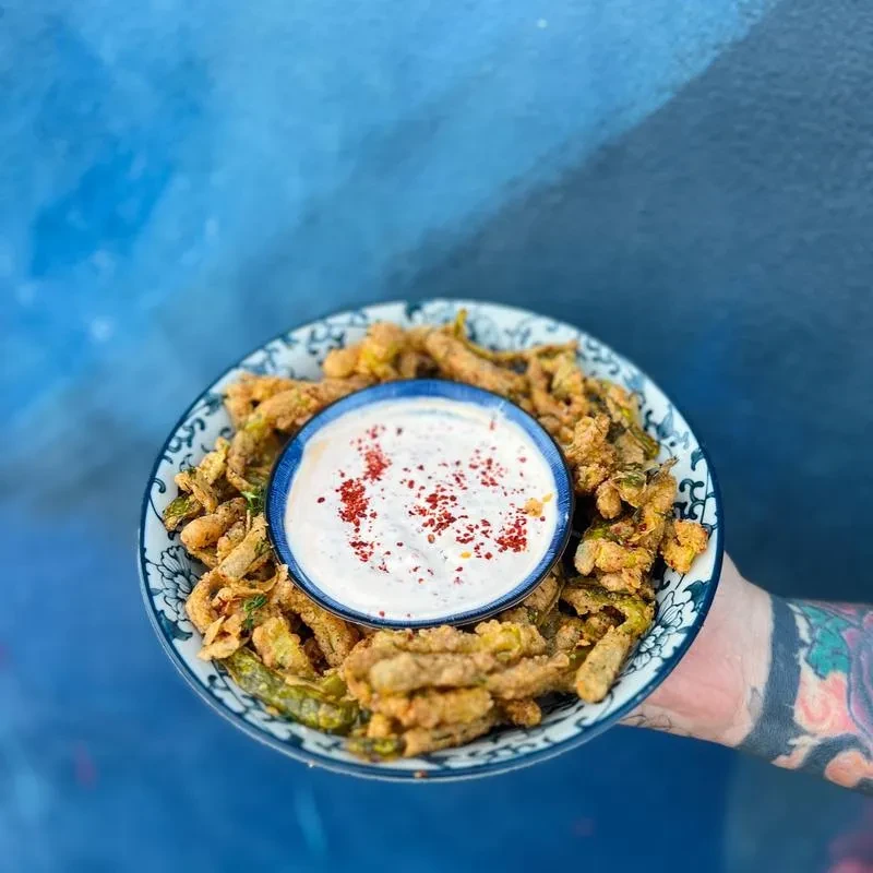 Fried green beans with red-flaked dip, held by a tattooed hand.