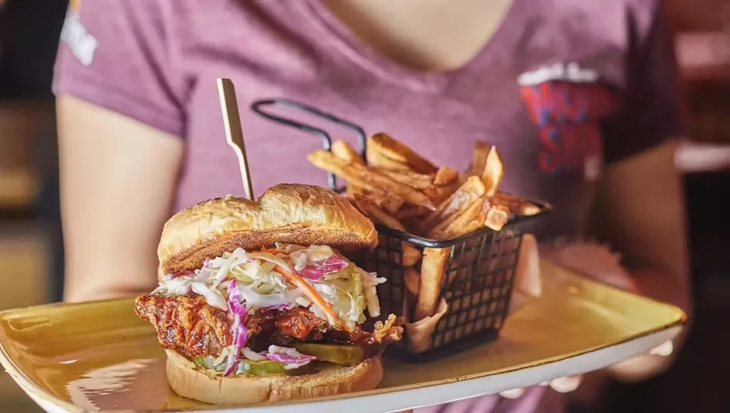 Person holding large sandwich with colorful coleslaw, pickles, and fries.