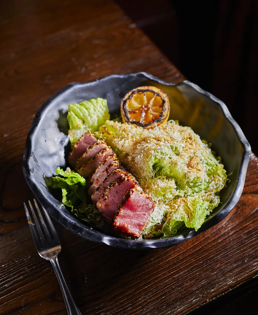 Seared tuna salad in dark bowl with lettuce, crumble, and grilled lemon.