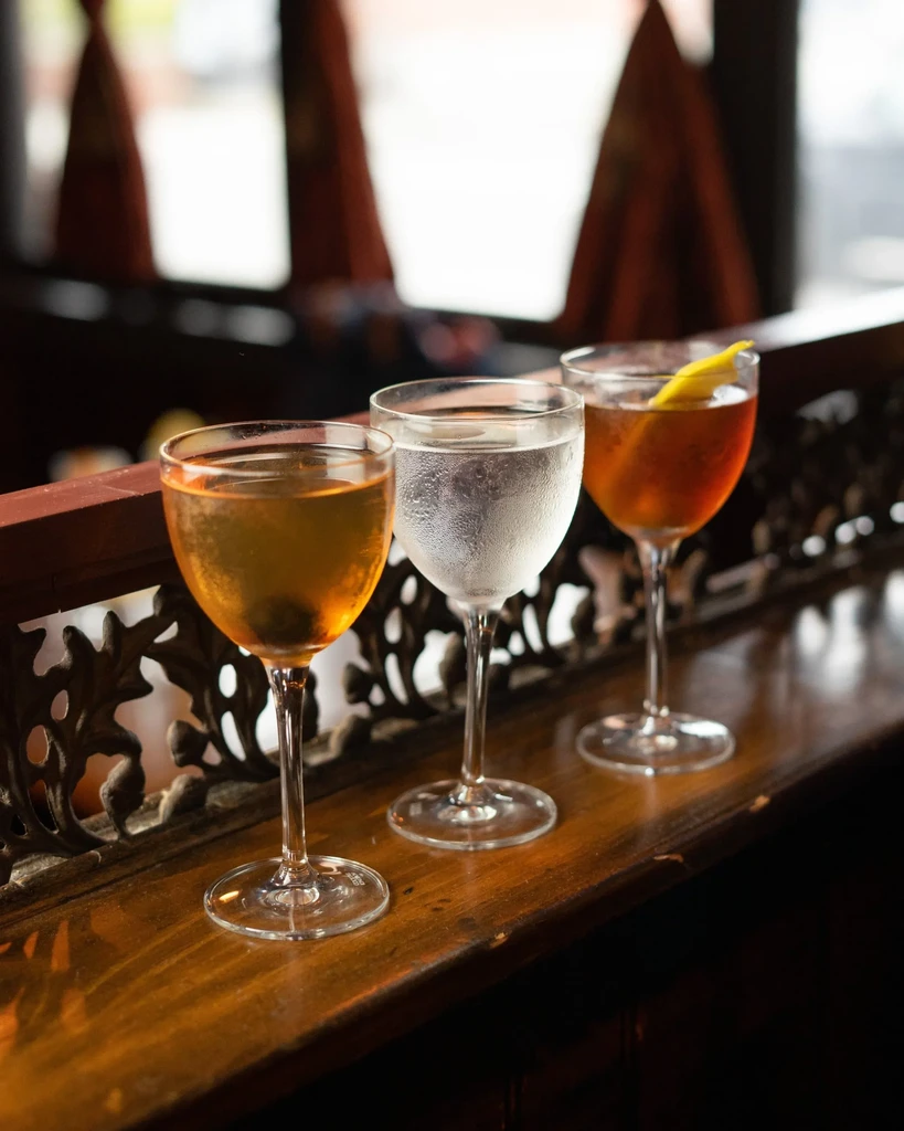 Three varied drinks in stemmed glasses on a wooden bar with railing.