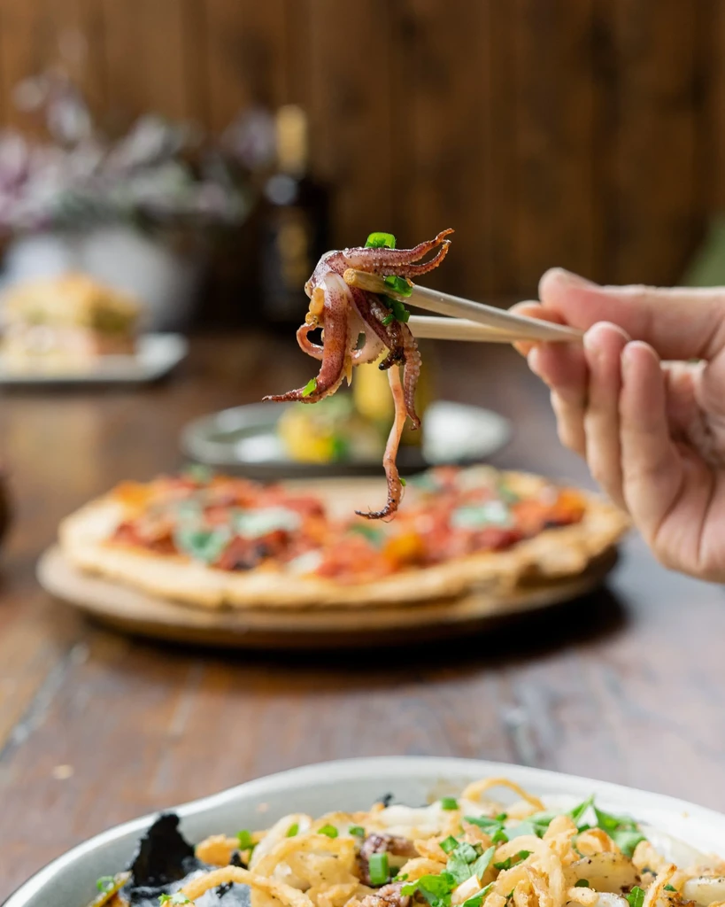 Chopsticks holding grilled squid with green onions; blurred pizza behind.