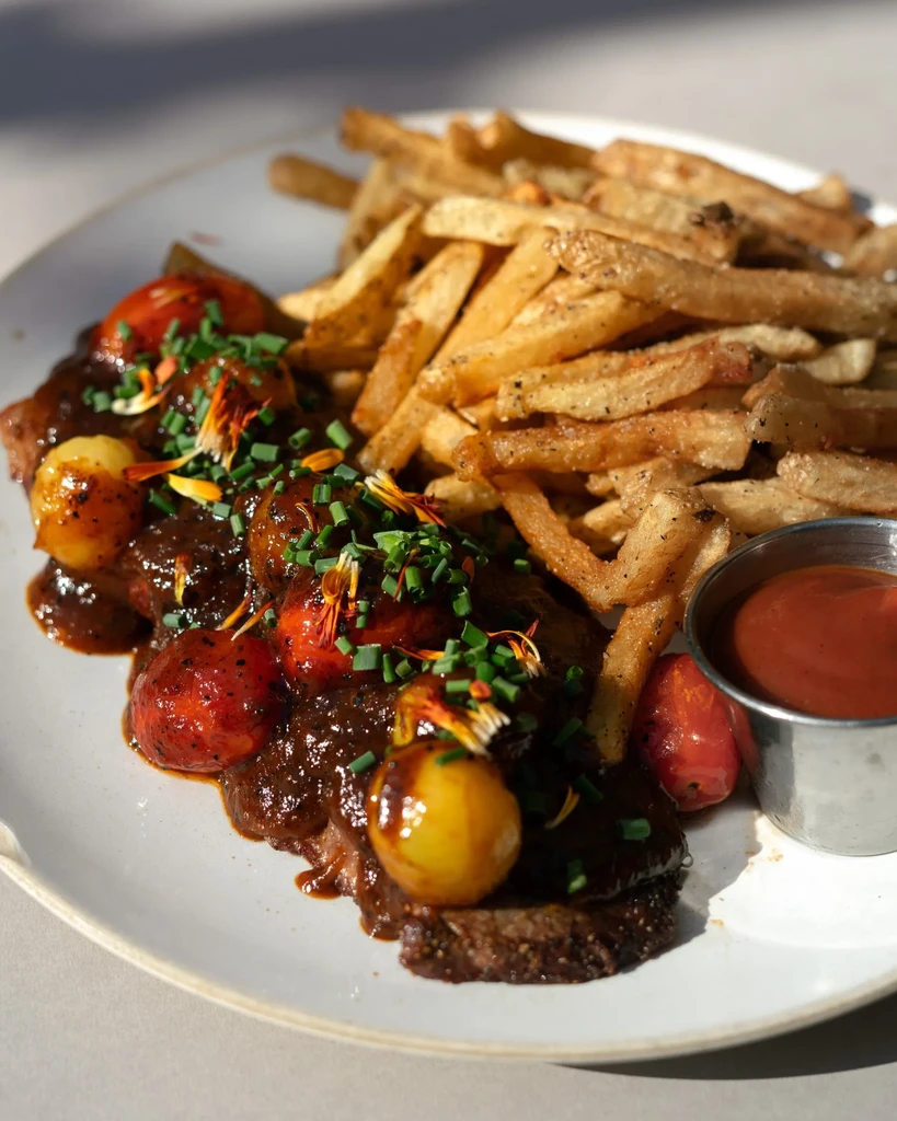 Close-up of meat with gravy, cherry tomatoes, chives, fries, and ketchup.