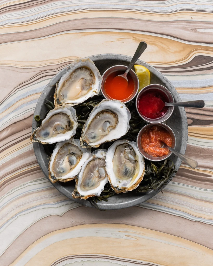 Platter of six shucked oysters, sauces, lemon, and green seaweed.