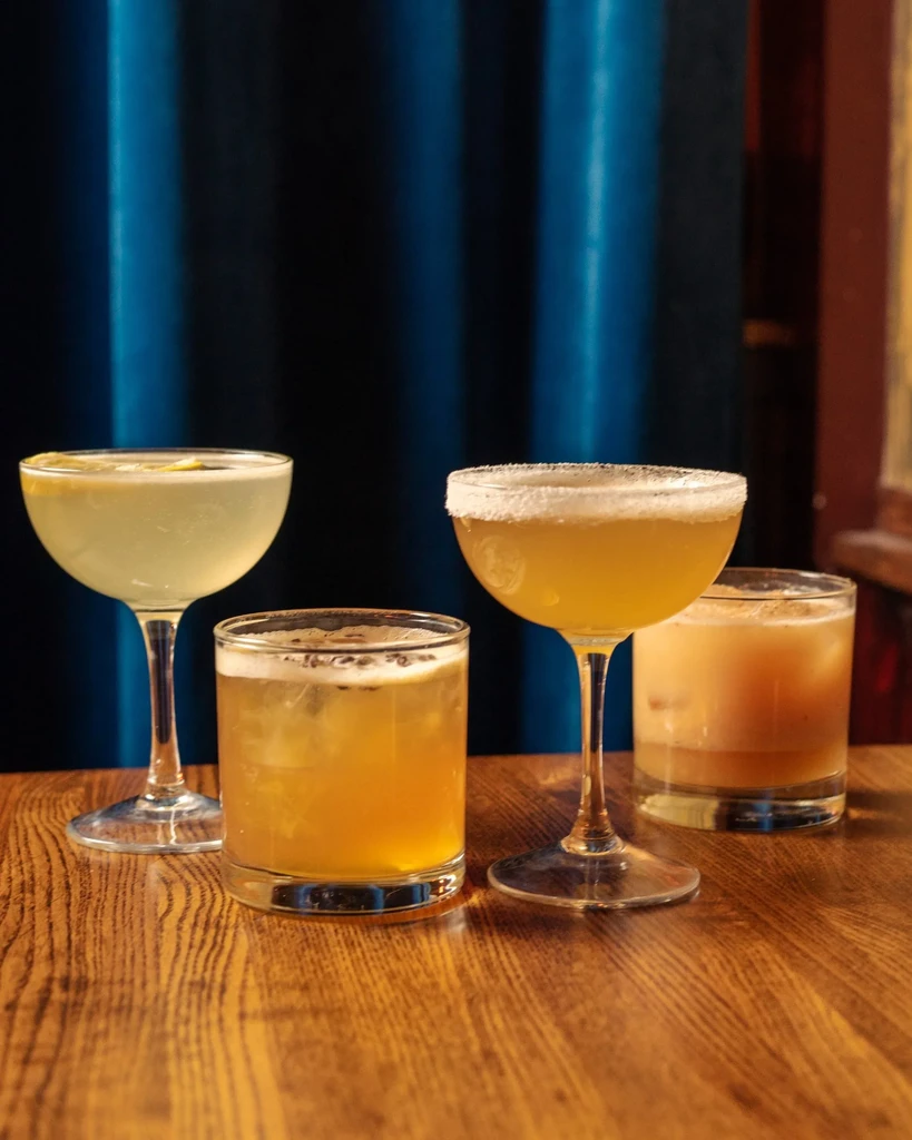 Four distinct cocktails on a wooden table against a dark blue curtain.