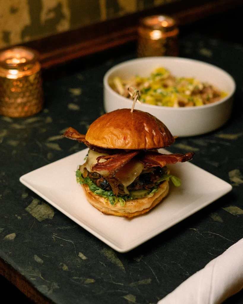 Gourmet burger with bacon, cheese, lettuce, and pickles on a dark table.