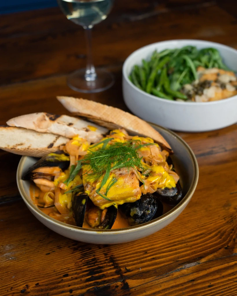 A bowl of seafood with mussels, fish, creamy sauce, dill, and bread.