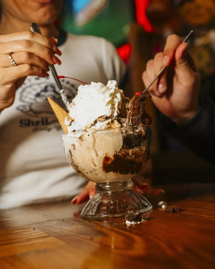 Two hands sharing a large sundae with ice cream, brownie, and cherry.