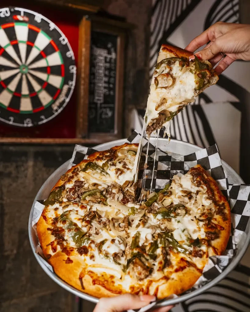 Hand lifting a slice of meat and vegetable pizza with a cheese pull.