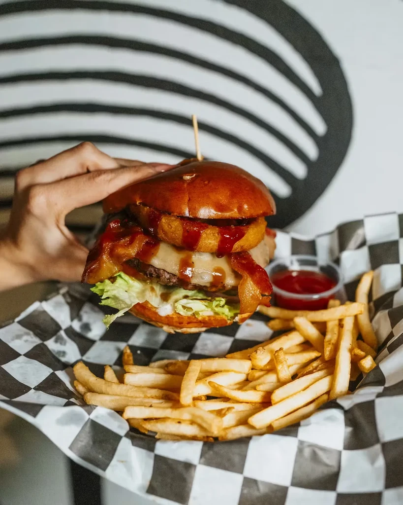 A hand holds a gourmet burger with bacon, onion rings, and fries.