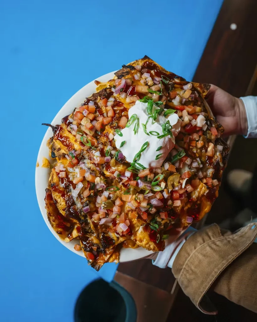 Hand holding loaded nachos with cheese, salsa, sour cream against blue background.