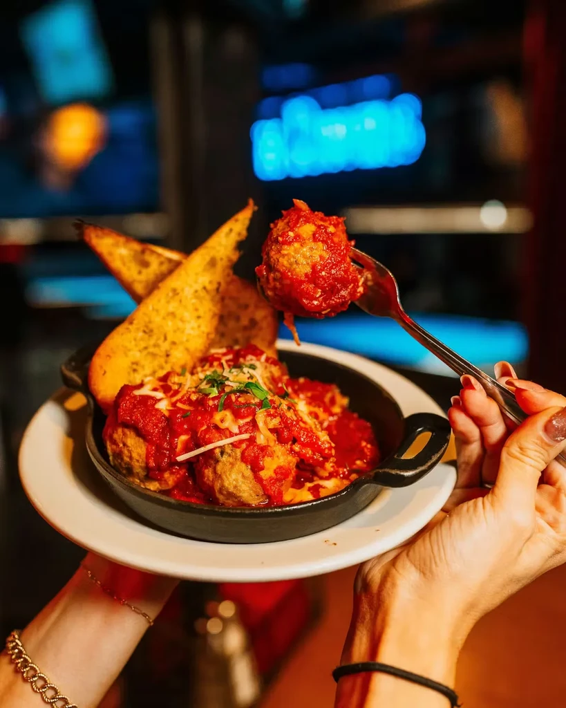 Meatballs in skillet with cheese, herbs, garlic bread, held with a fork.