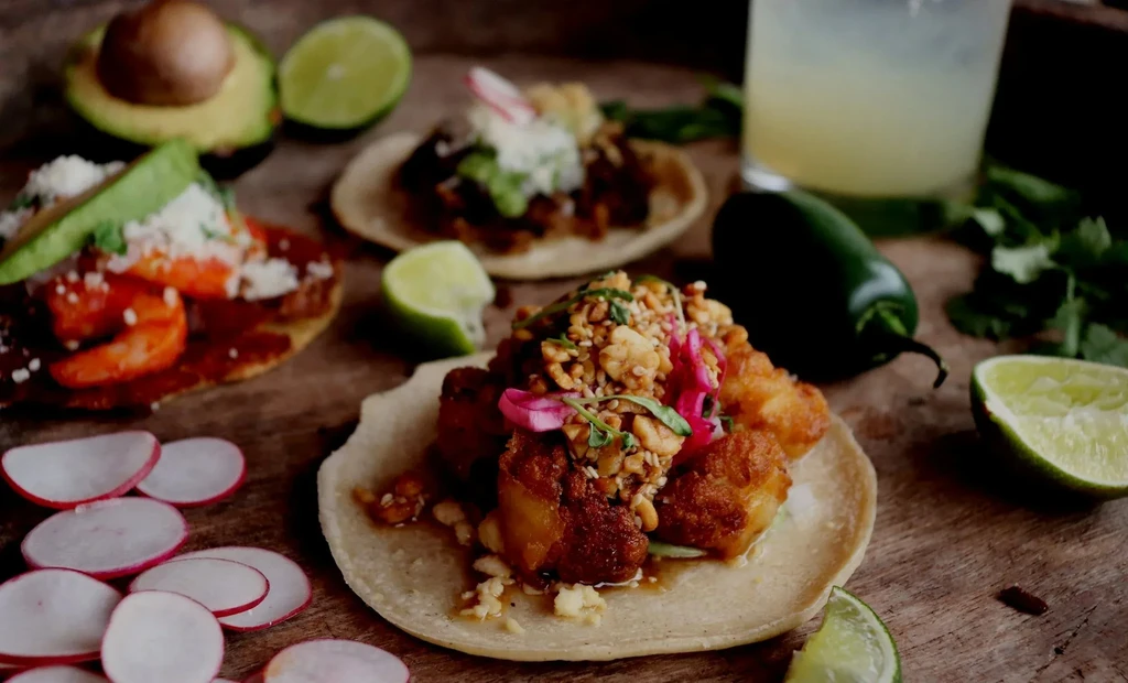 Several fish and shrimp tacos with fresh garnishes on a wooden surface.