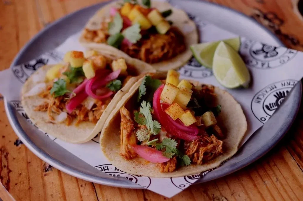 Three colorful meat tacos with pineapple, onions, cilantro on wood table.