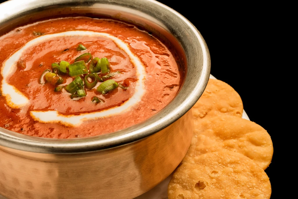 Vibrant reddish-orange Indian curry in copper bowl with flatbreads on black background.