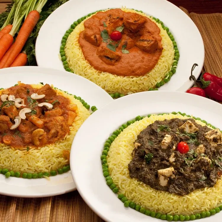 Three plated Indian curries with rice, peas, carrots, and chilies.