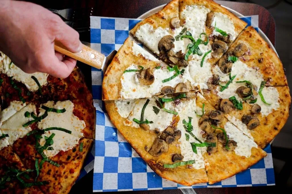 Hand lifts slice of white pizza with mushrooms and herbs.