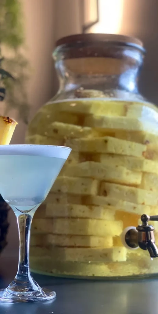 Frothy cocktail with pineapple beside a pineapple-infused drink dispenser.