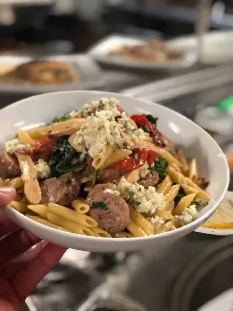 Close-up of hand holding penne pasta with sausage, blue cheese, and vegetables.