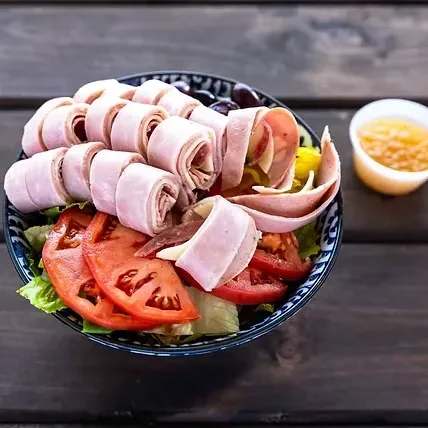 Generous salad with ham, tomatoes, olives, and dressing in blue bowl.