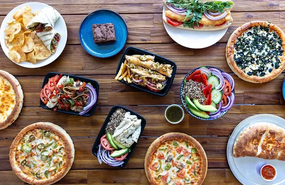 Overhead view of diverse pizzas, salads, and sandwiches on a wooden table.
