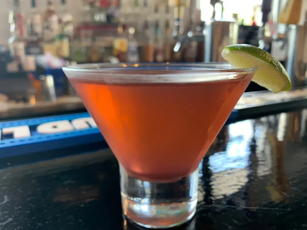 Red-orange cocktail in martini glass with lime on bar.