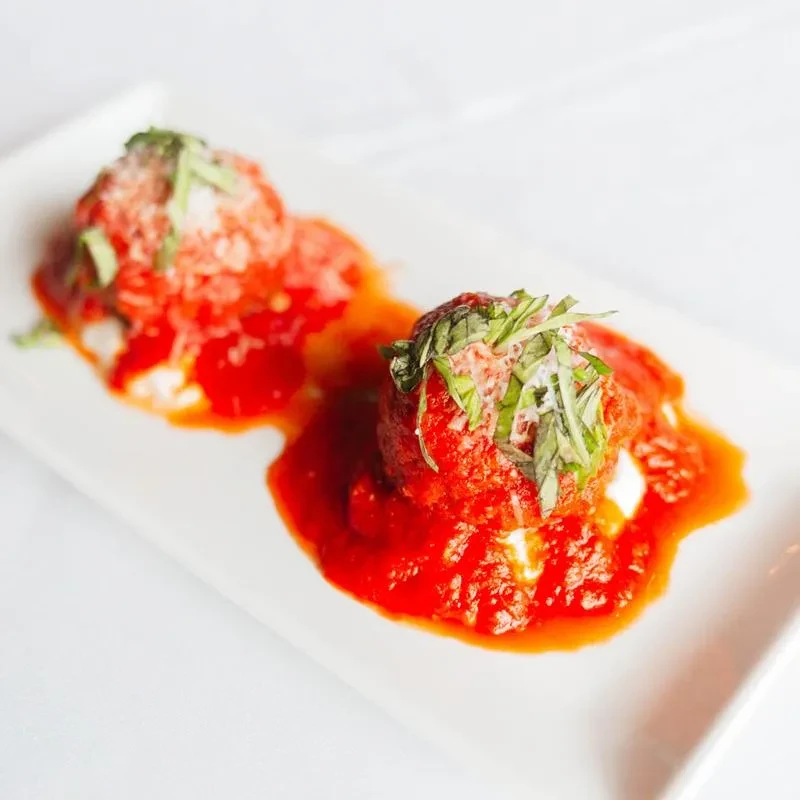 Two meatballs with red tomato sauce, parmesan, and basil on white plate.