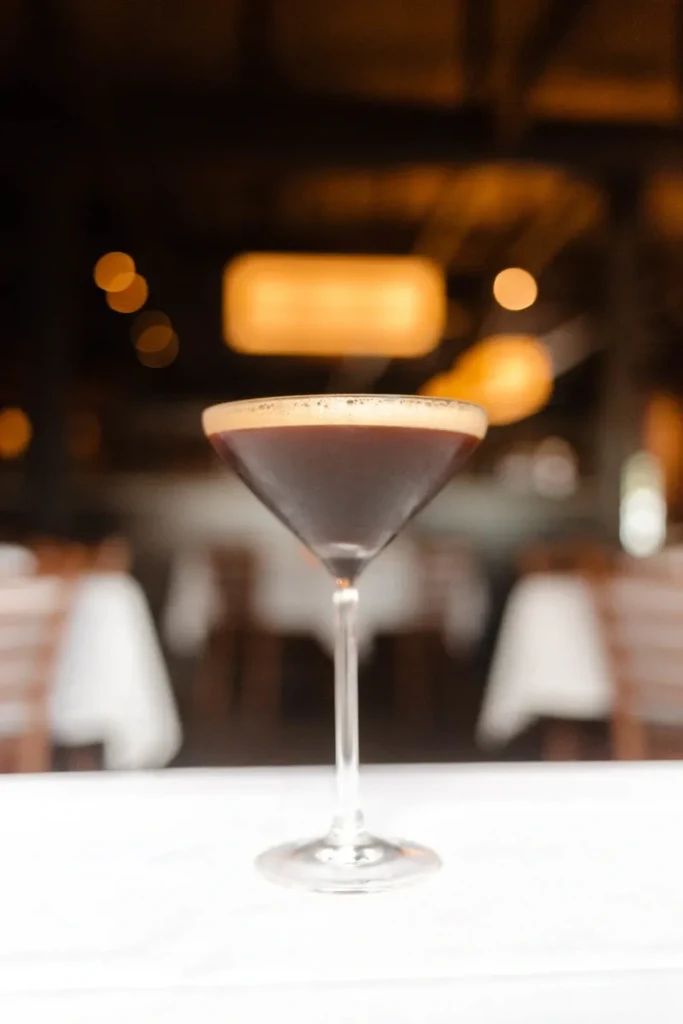 Dark cocktail with foam in martini glass, blurred bar background.
