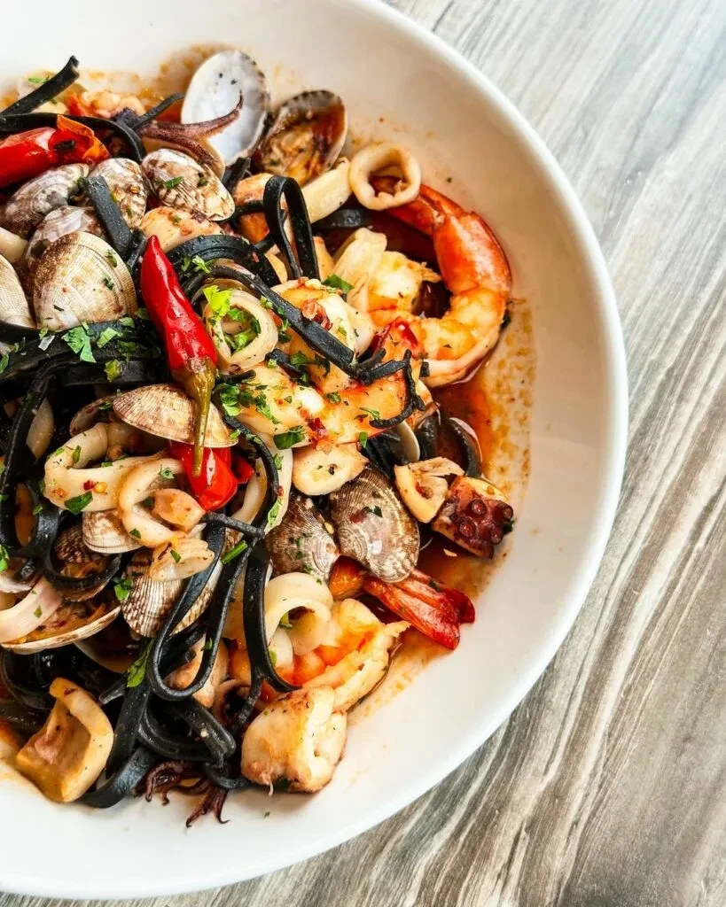 Top-down black squid ink pasta with seafood, red sauce, chili, and herbs.