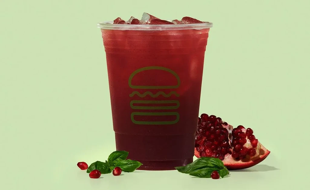 Red drink in logo cup, with ice, pomegranate, and green leaves.