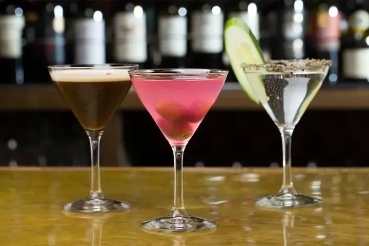 Three cocktails: dark, pink with garnishes, clear with cucumber, on a bar.
