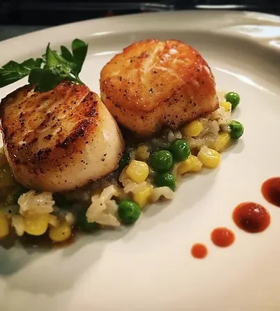 Two golden-brown pan-seared scallops on rice, corn, peas, and sauce.
